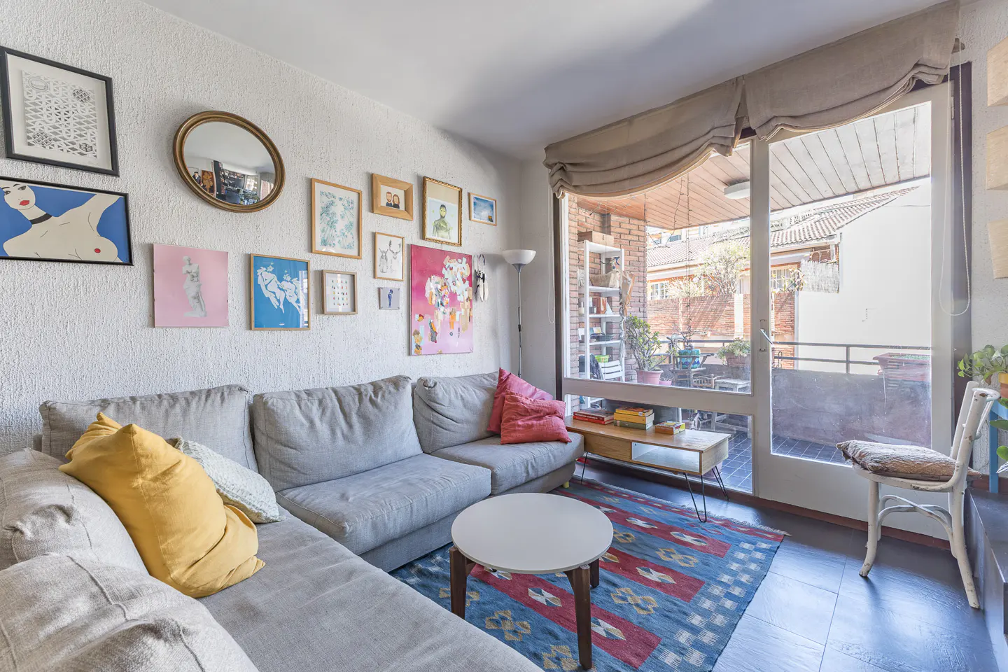 Living room with a gray sectional sofa, colorful art on the wall, and a large window leading to a balcony.