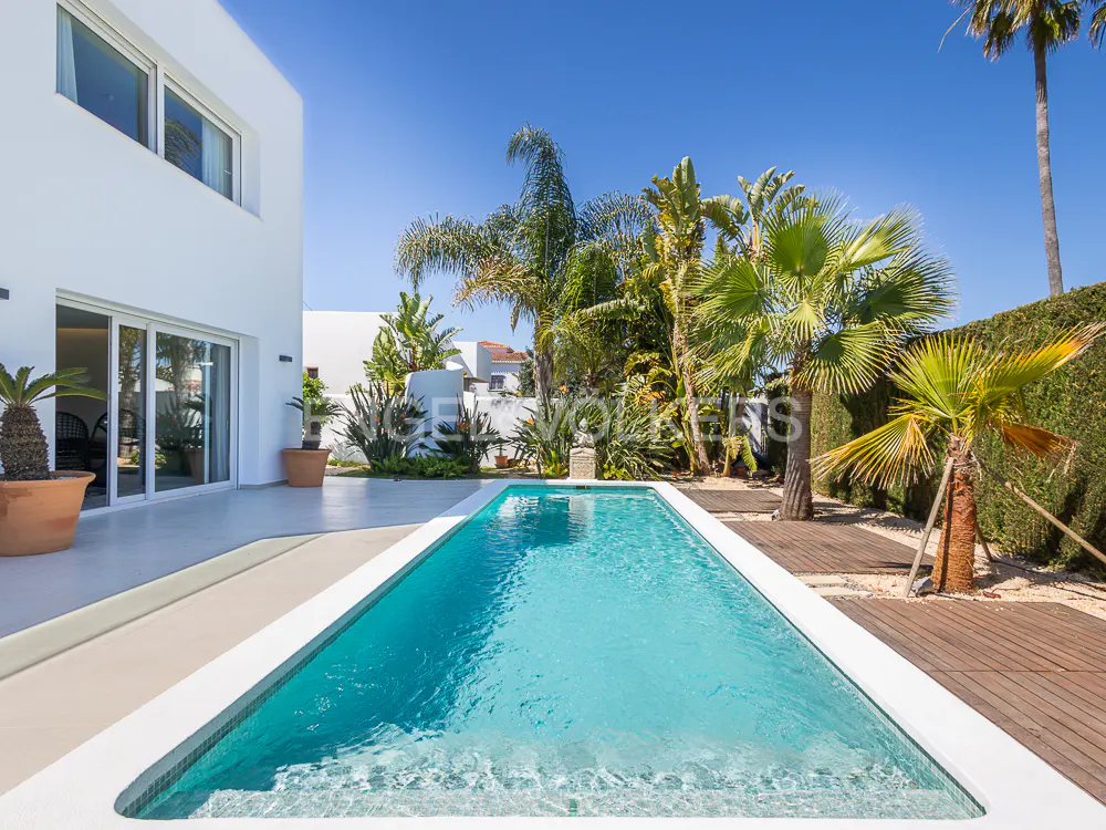 A modern white house with a rectangular pool, palm trees, and a clear blue sky.