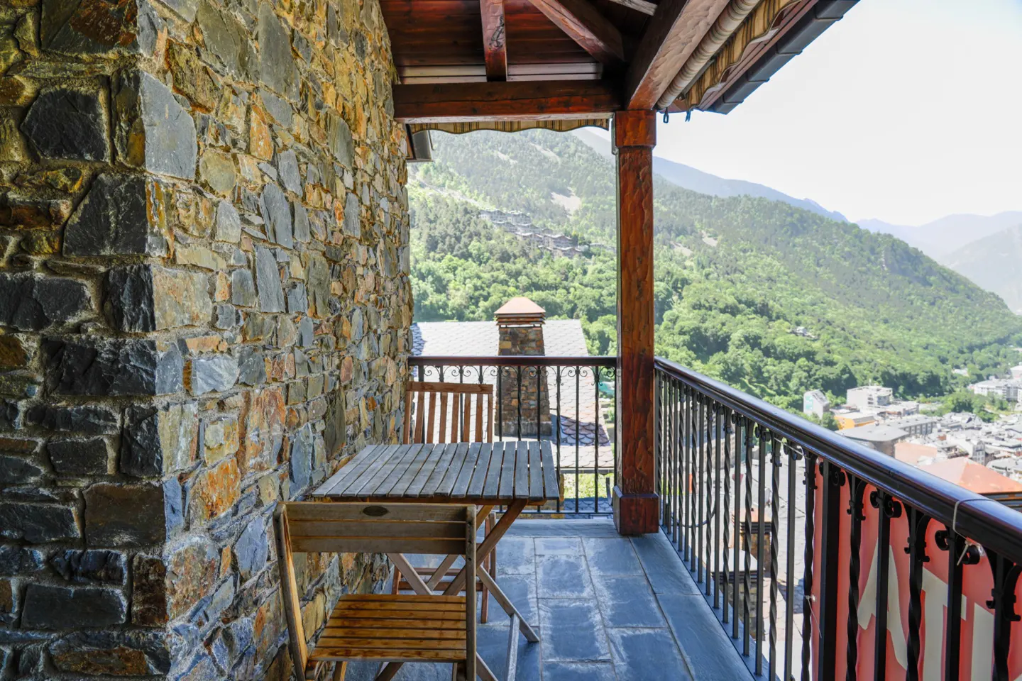 Balcony with stone wall, wooden table and chairs, and a view of green mountains and a town below.