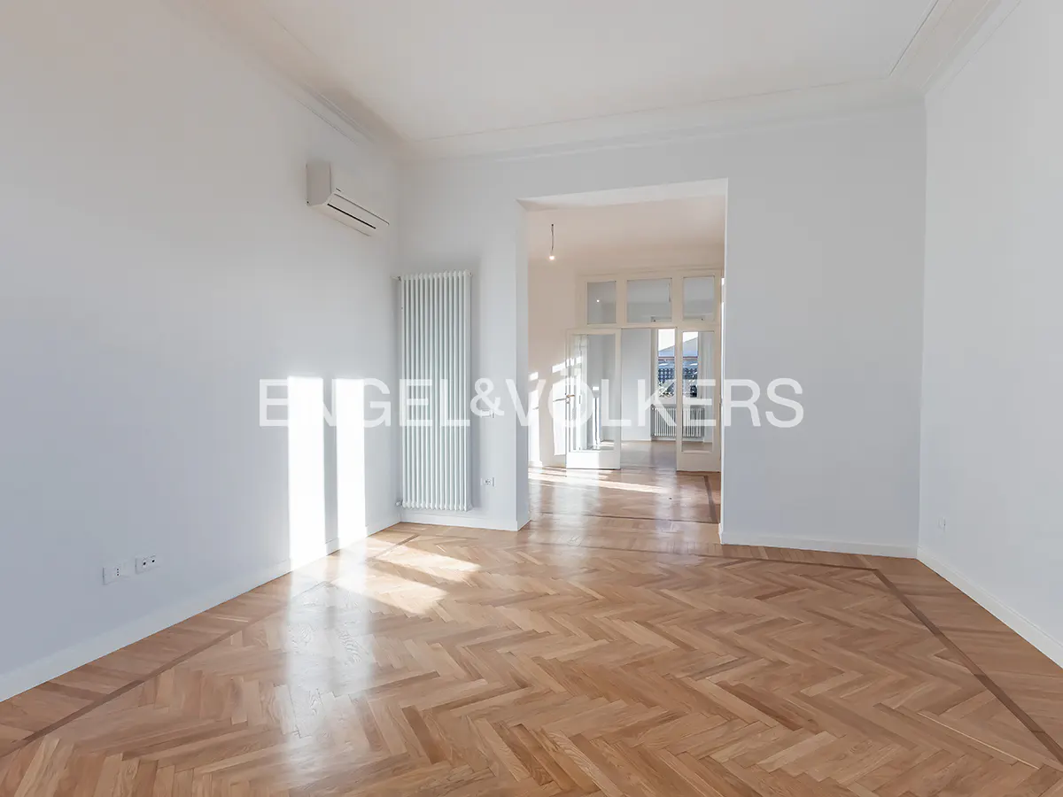Bright, empty room with herringbone wood floors, white walls, radiator, and doorway to another room.
