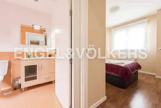 View through a doorway showing a bathroom with peach tiles and a bedroom with a bed featuring a dark red blanket.
