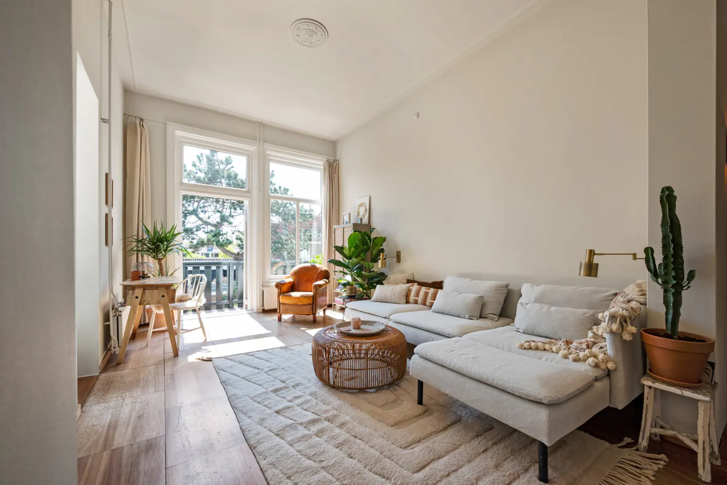 Bright living room with a gray sectional sofa, rattan coffee table, and a large window overlooking a balcony. A cactus sits on a stool.