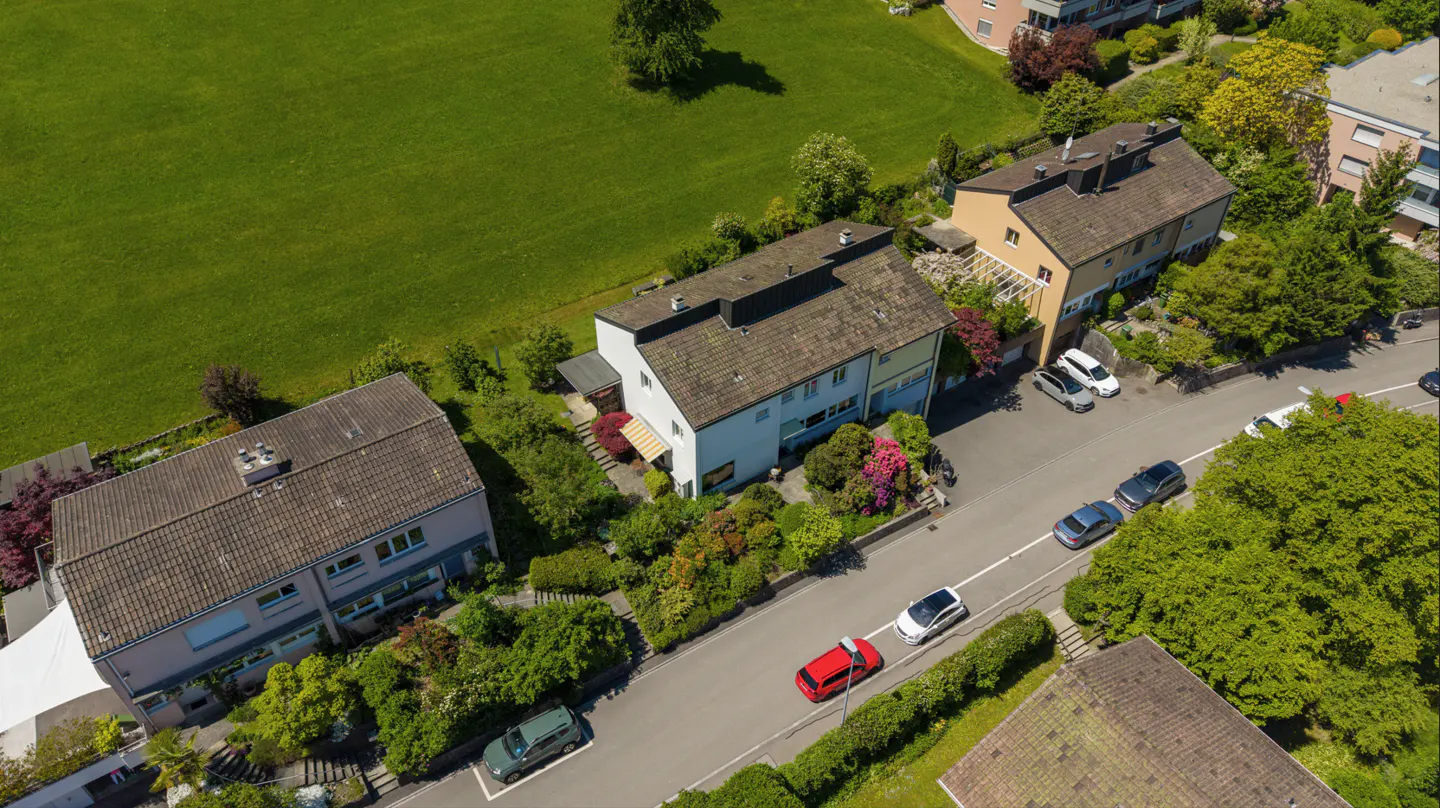 Aerial view of houses with cars parked on the street, surrounded by green lawns and trees.
