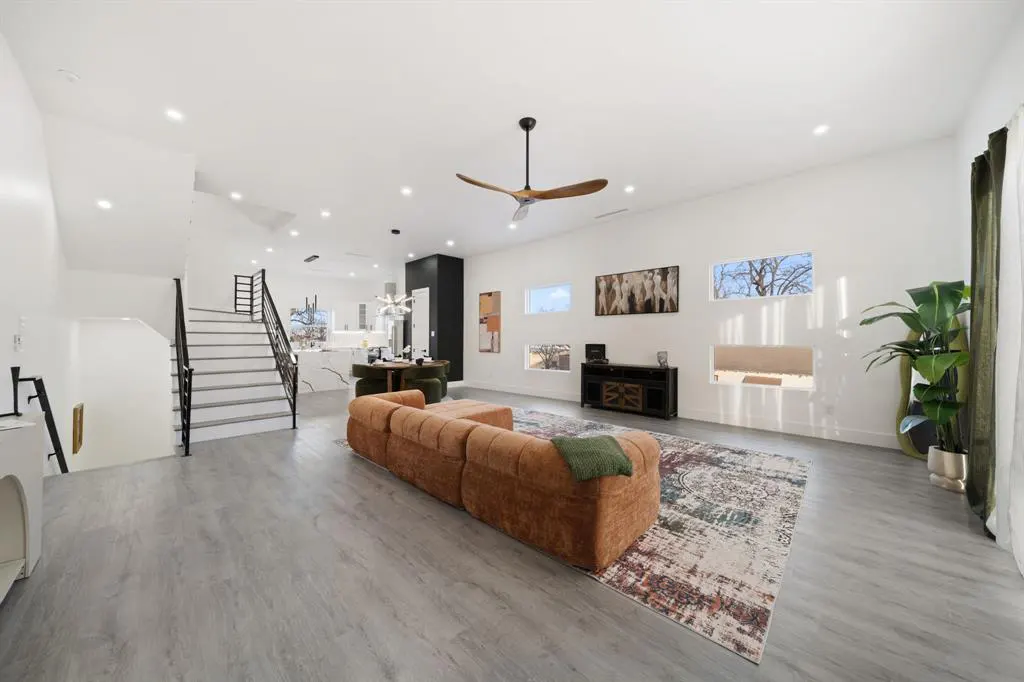 Bright, modern living room with gray floors, white walls, and a brown sectional sofa on a patterned rug. Stairs and dining area visible.
