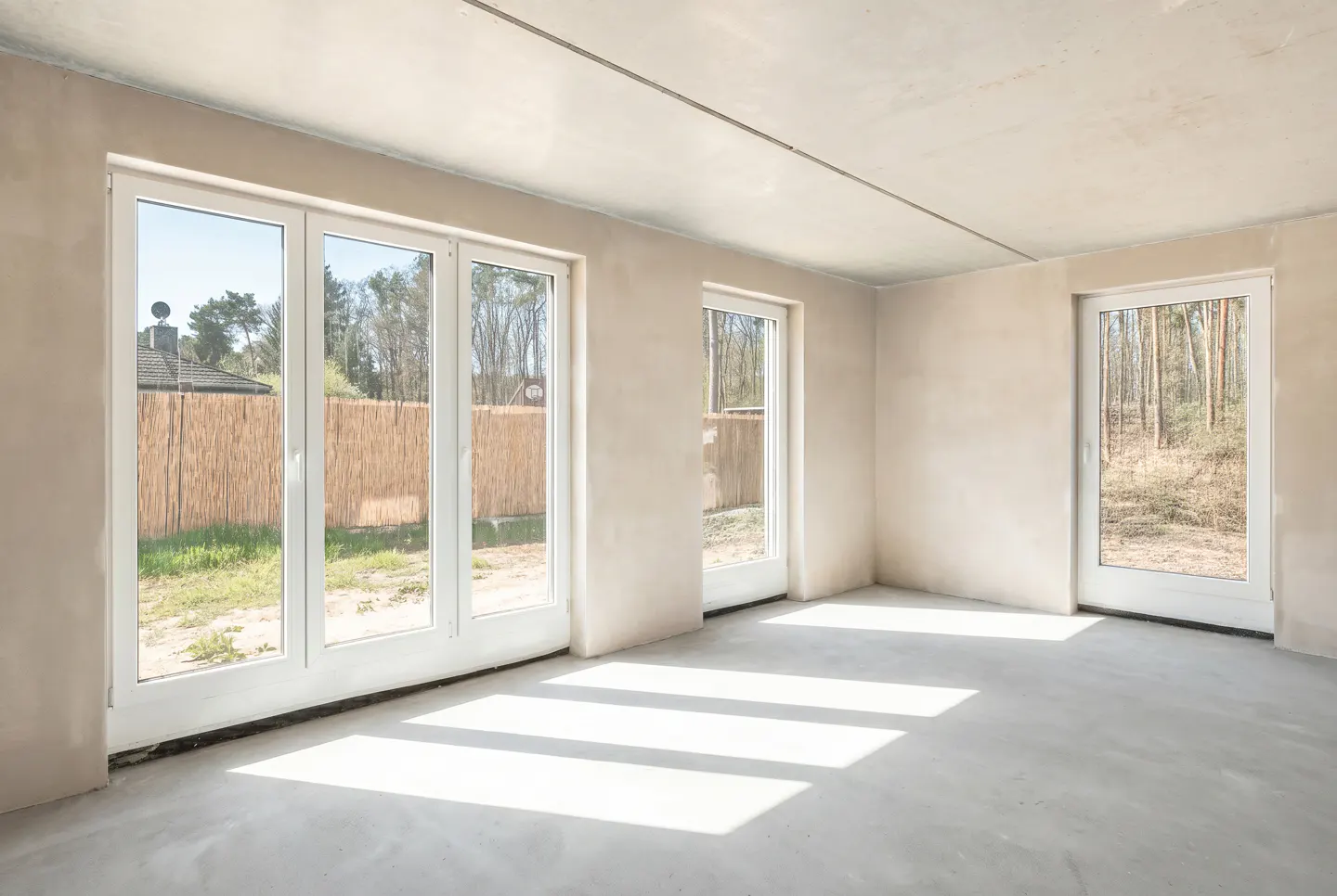 Unfinished room with three white-framed windows, bare walls, and concrete floor with sunlight streaming in.