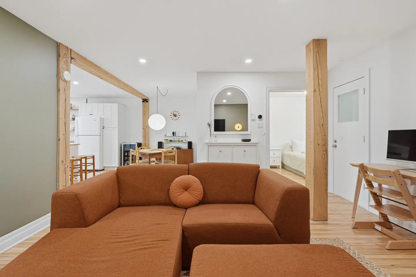Interior view of a modern living room with a brown sectional sofa, wooden beams, and white walls. A kitchen and dining area are visible in the background.