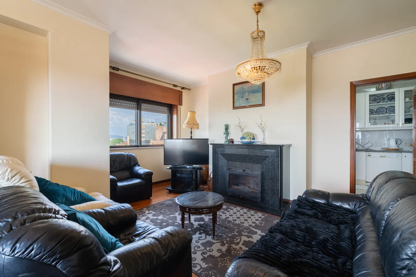 Living room with black leather sofas, armchair, TV, and fireplace. A chandelier hangs from the ceiling. A kitchen is visible through a doorway.