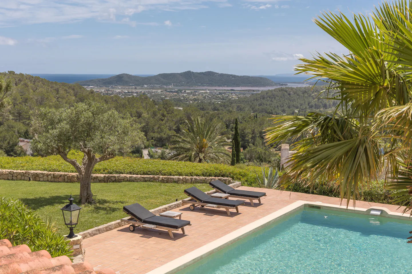 Outdoor pool with three lounge chairs on a brick patio, overlooking green hills and the ocean under a blue sky.