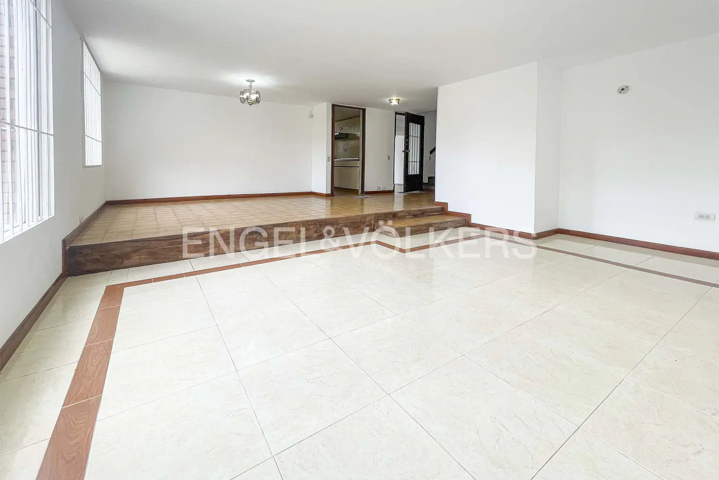 Bright, empty living room with white walls and beige tile floor. A raised platform is in the back. A doorway leads to a kitchen and stairs.