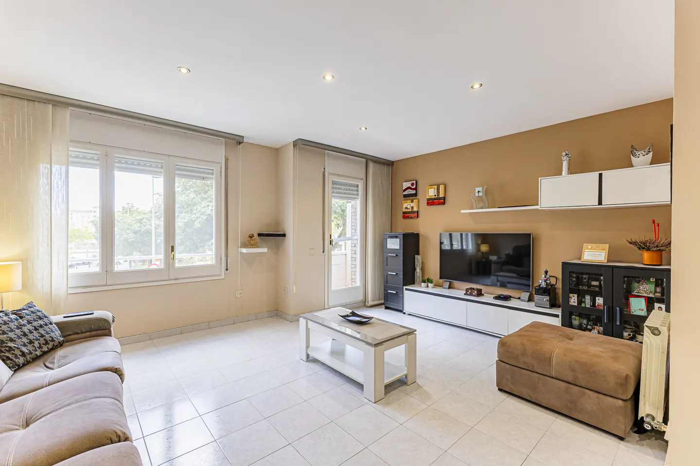 Living room with brown sofa, white coffee table, TV, and white tile floor. Natural light from windows.