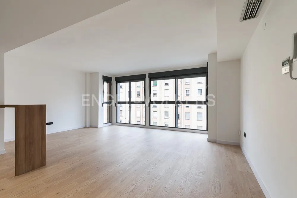 Empty, bright living room with light wood floors, white walls, and large windows overlooking a city building.