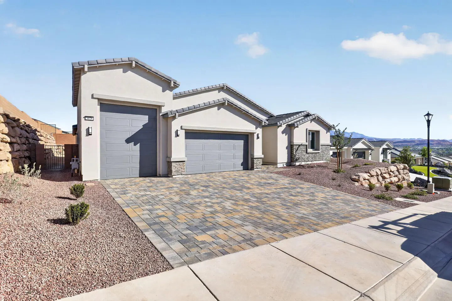 A modern, single-story home with two gray garage doors, a patterned brick driveway, and desert landscaping under a blue sky.
