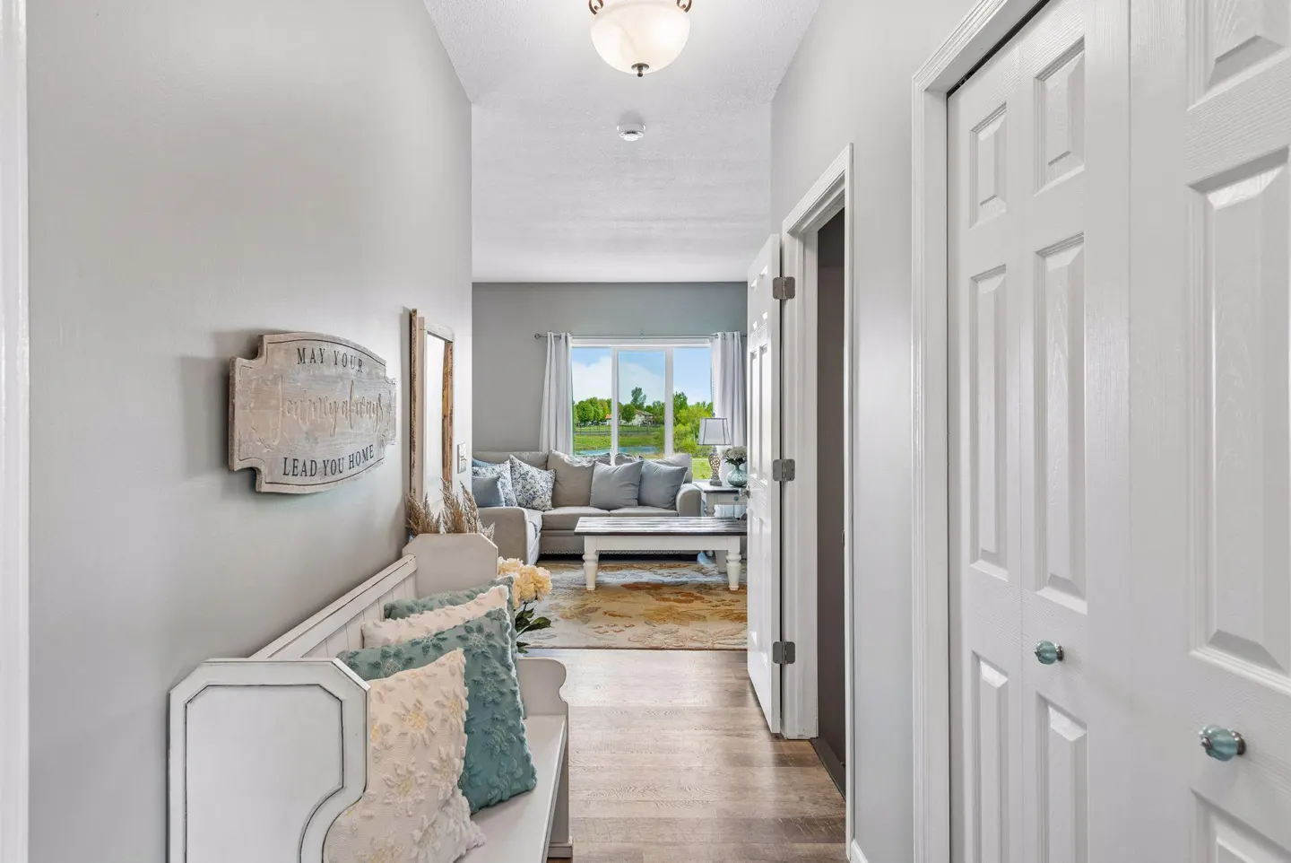 Hallway view with a white bench, decorative pillows, and a sign on the wall. A living room is visible in the background.