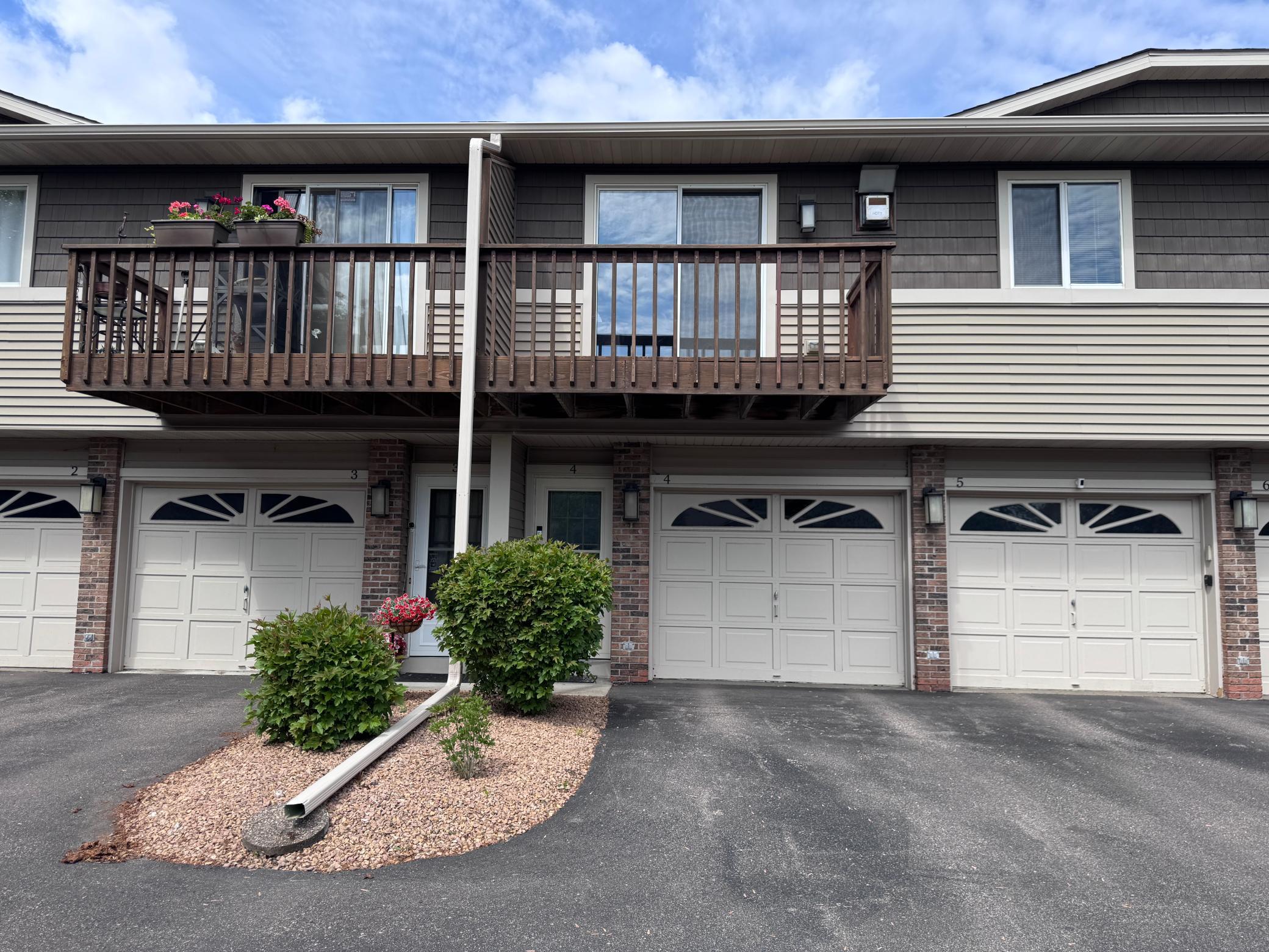 A two-story townhouse with brown siding, white garage doors, and a wooden balcony.