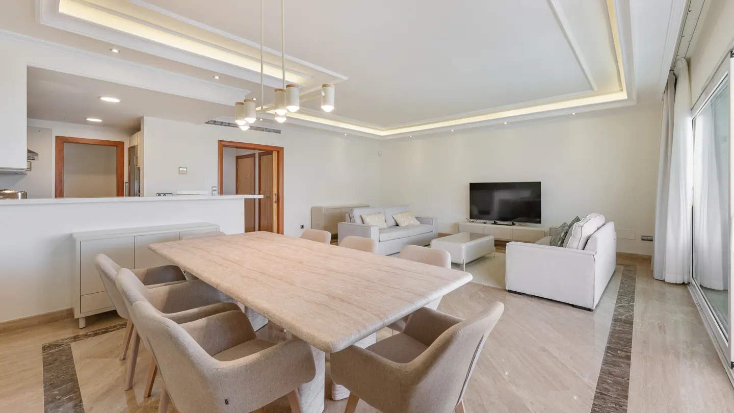 Modern open-plan living room with a light wood dining table, beige armchairs, white sofas, and a wall-mounted TV.