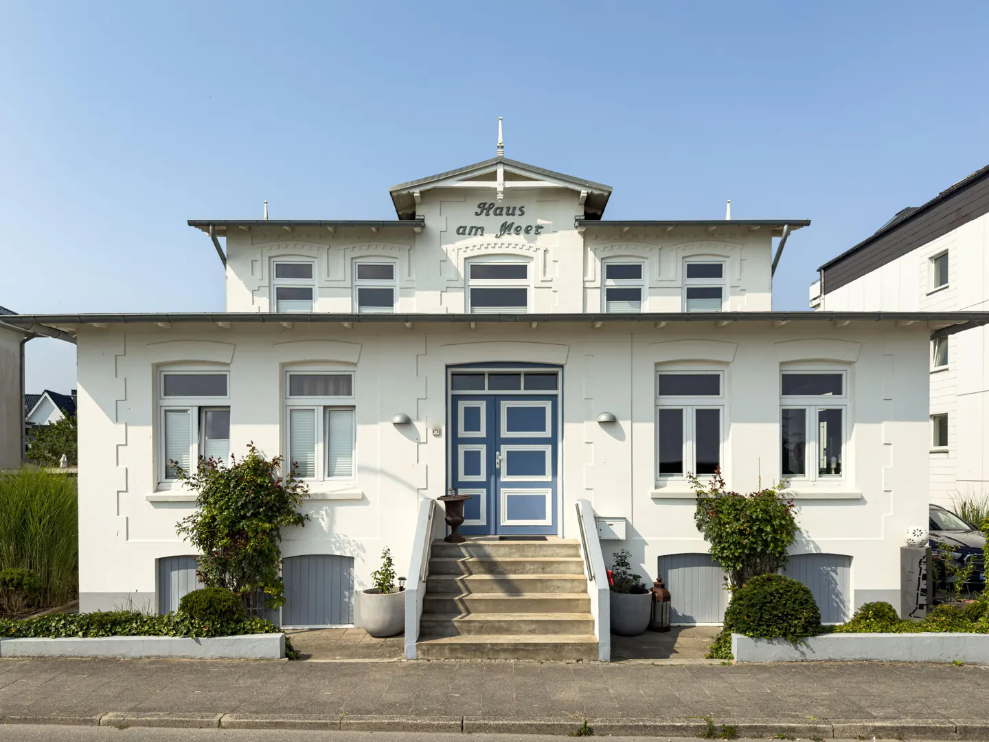 Two-story white house with a blue door and "Haus am Meer" sign above the entrance. Steps lead to the door, with plants on either side.