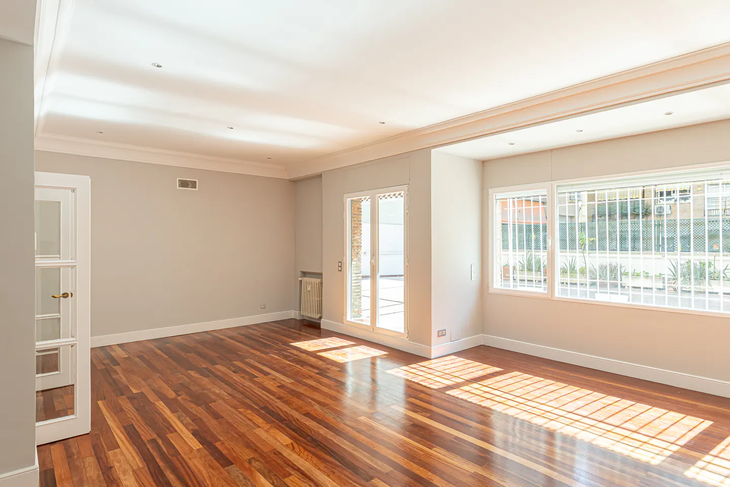 Bright, empty room with hardwood floors, white trim, and gray walls. Sunlight streams through barred windows and glass doors.