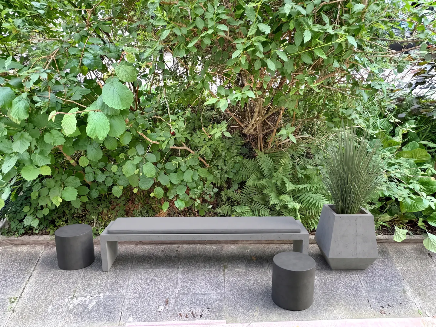 Modern gray bench with two matching stools and a tall planter on a stone patio in front of lush green garden foliage.