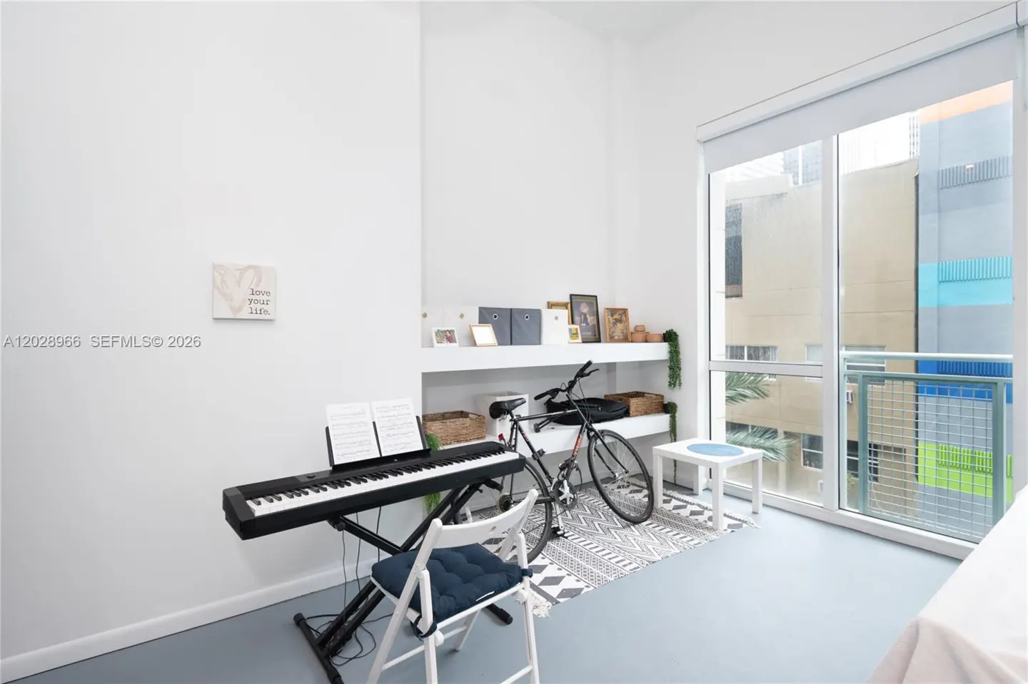 Modern room with a digital piano, white chair, bicycle, and shelving unit, next to a large window with city views.