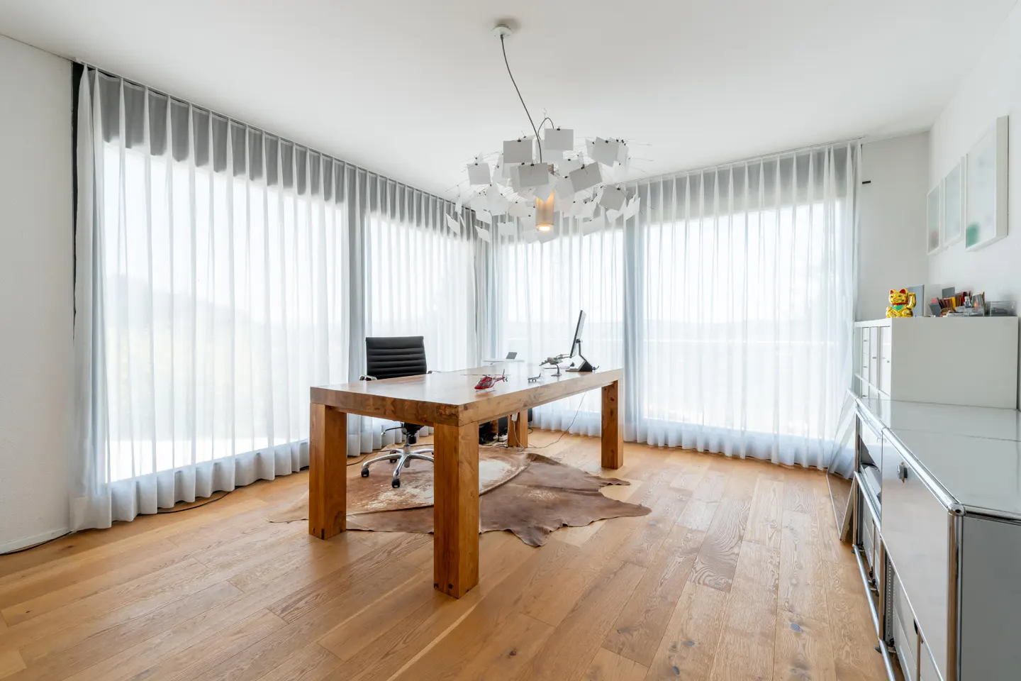 Bright home office with wood floors, a large wood desk, black chair, and white curtains. A cowhide rug sits under the desk.