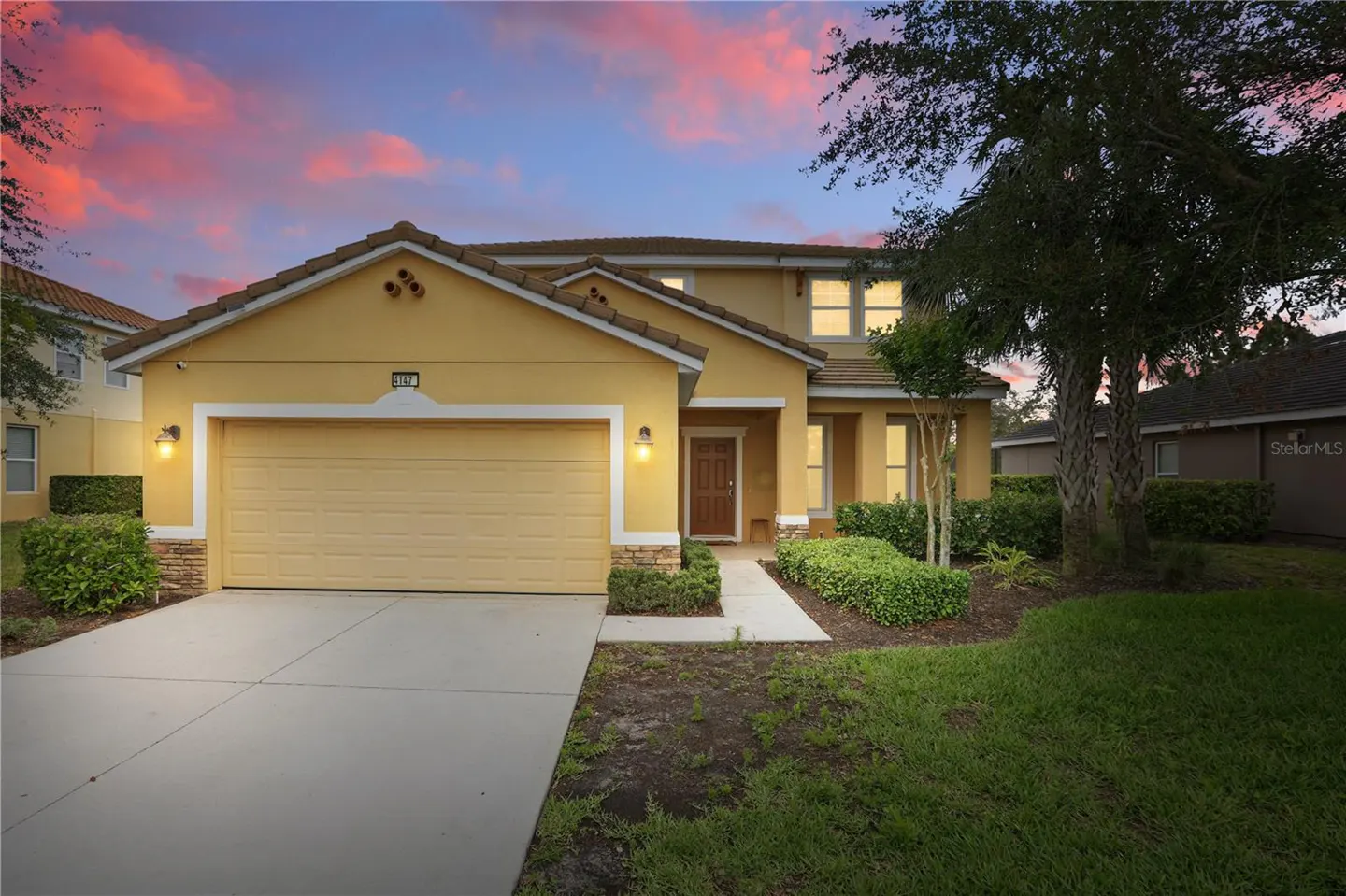 Two-story yellow house with a brown roof, a two-car garage, and a green lawn at dusk.