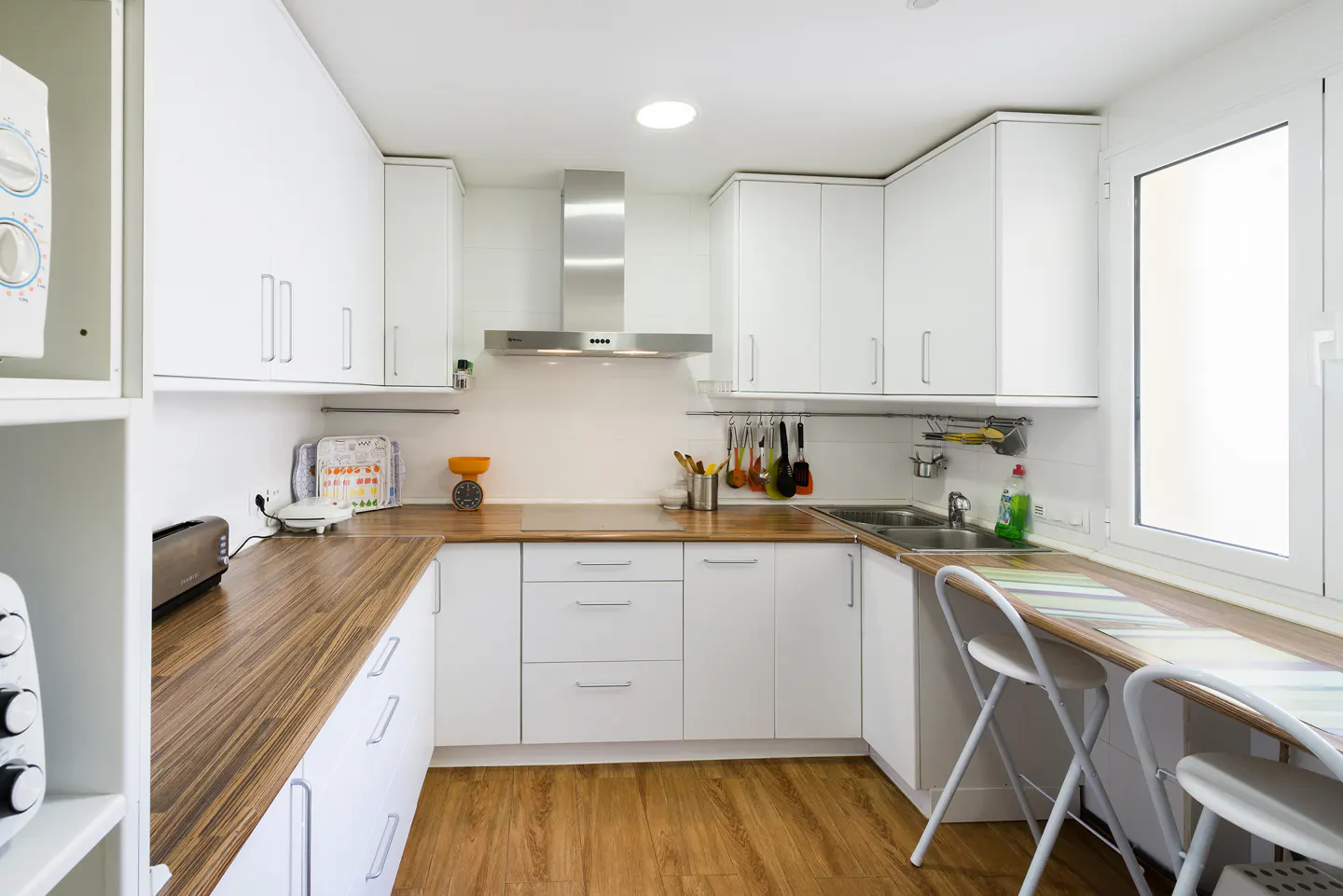 Bright kitchen with white cabinets, wood countertops, and wood floor. Stainless steel range hood and sink. Two white folding chairs at a counter.