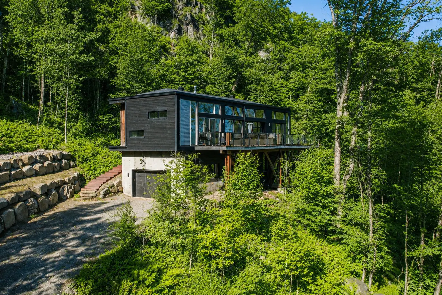 Modern black house with glass walls, nestled in a lush green forest on a hillside. Stone retaining wall and driveway visible.