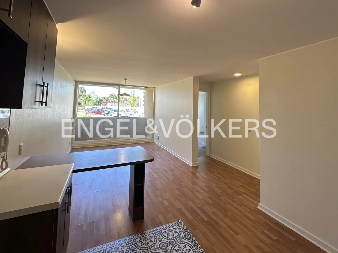 A bright apartment interior with wood floors, a kitchen island, and a large window overlooking a parking lot. The Engel & Völkers logo is superimposed.