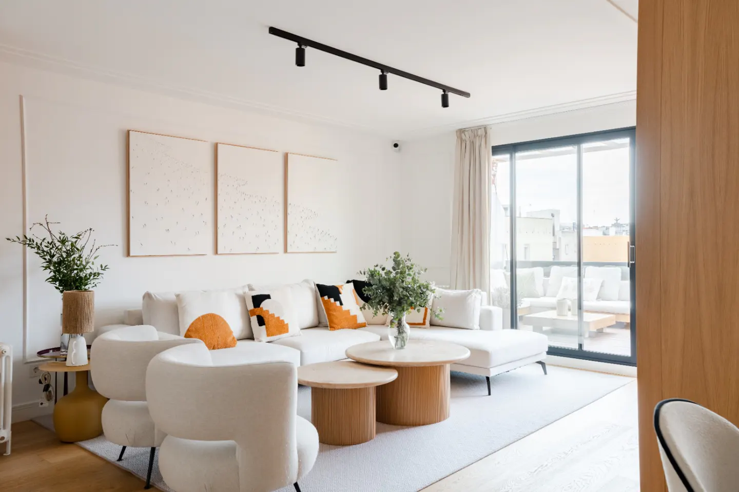 Bright living room with white sectional sofa, round wood tables, and abstract art. Balcony with outdoor seating visible through sliding glass doors.