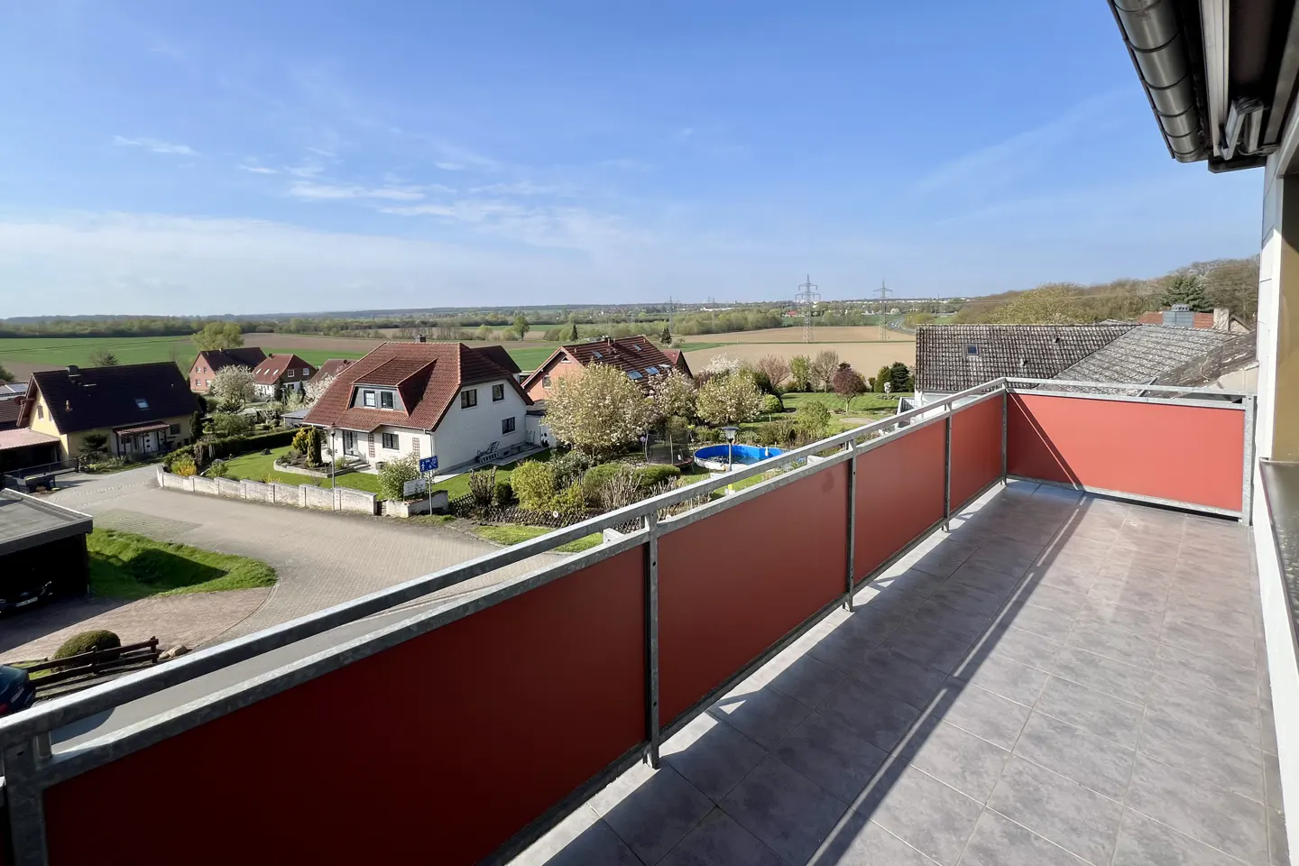 View from a balcony with a red railing overlooking houses, fields, and a blue sky.