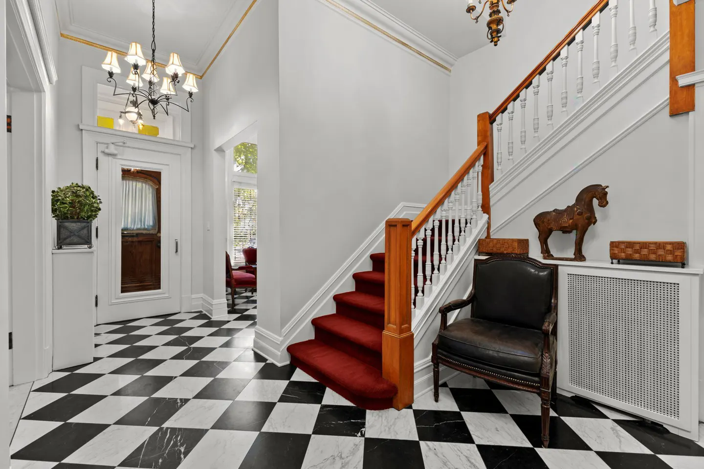Foyer with black and white checkered floors, white walls, staircase with red carpet, and a black leather chair.