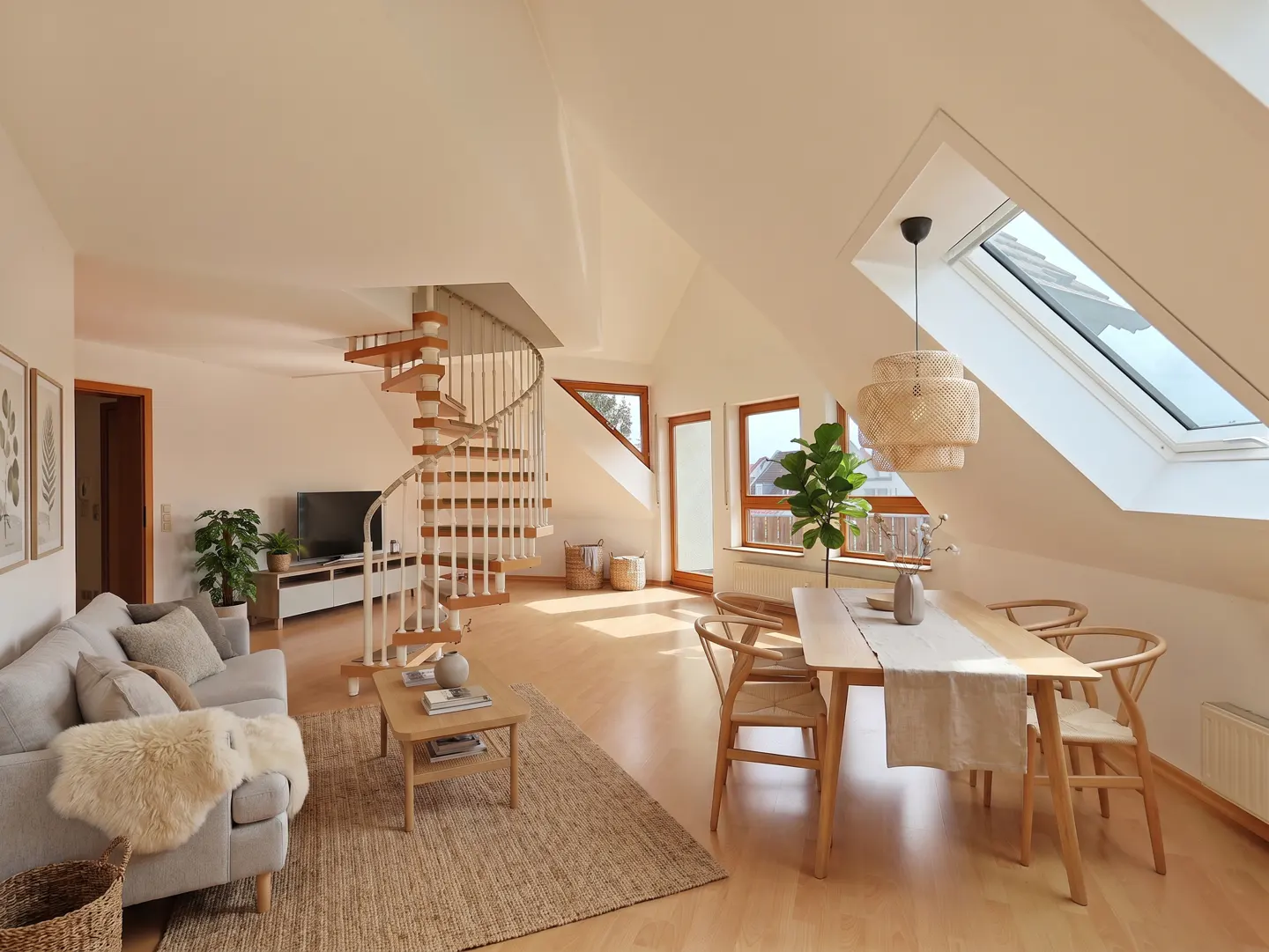 Bright, open-plan living space with a spiral staircase, light wood floors, and a dining area under a skylight. A gray sofa and TV are visible.