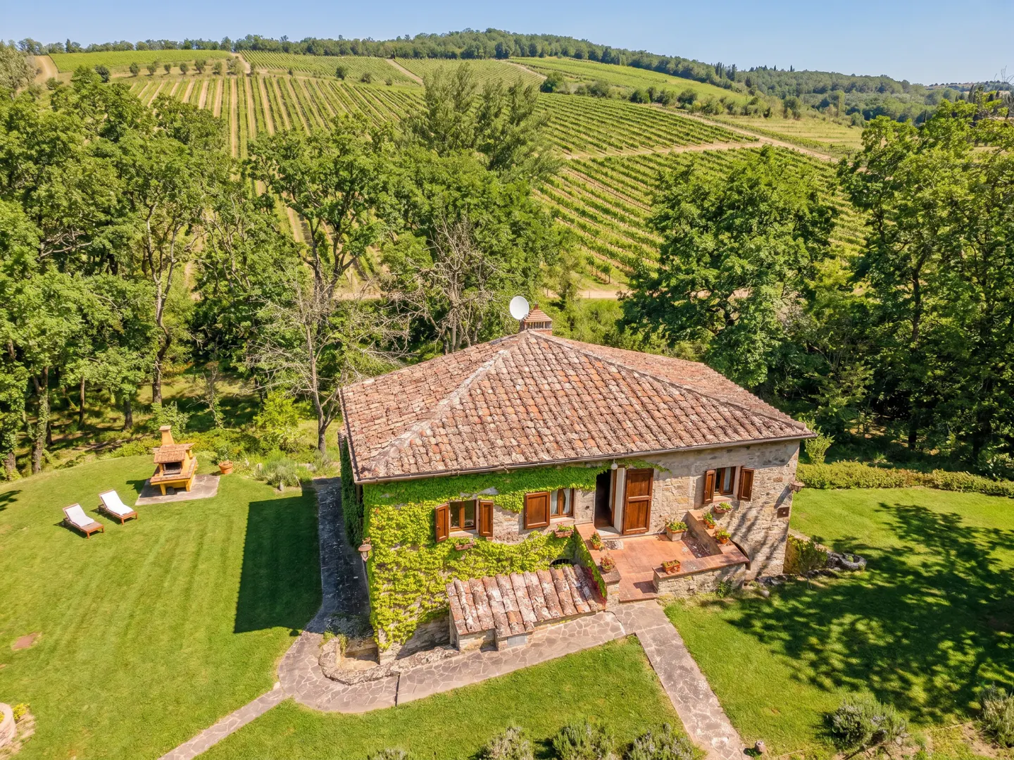 Aerial view of a stone house with a red tile roof, surrounded by green trees and a vineyard in the background.