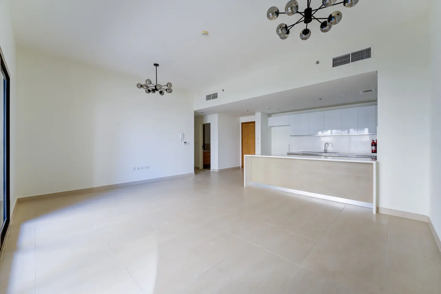 Bright, empty living room with white walls, tile floor, and open kitchen. Modern chandeliers hang from the ceiling.