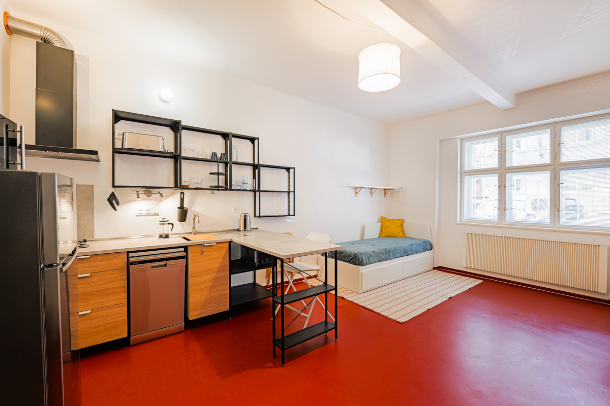 Studio apartment with a kitchen featuring wood cabinets, black shelving, and a daybed on a bright red floor.