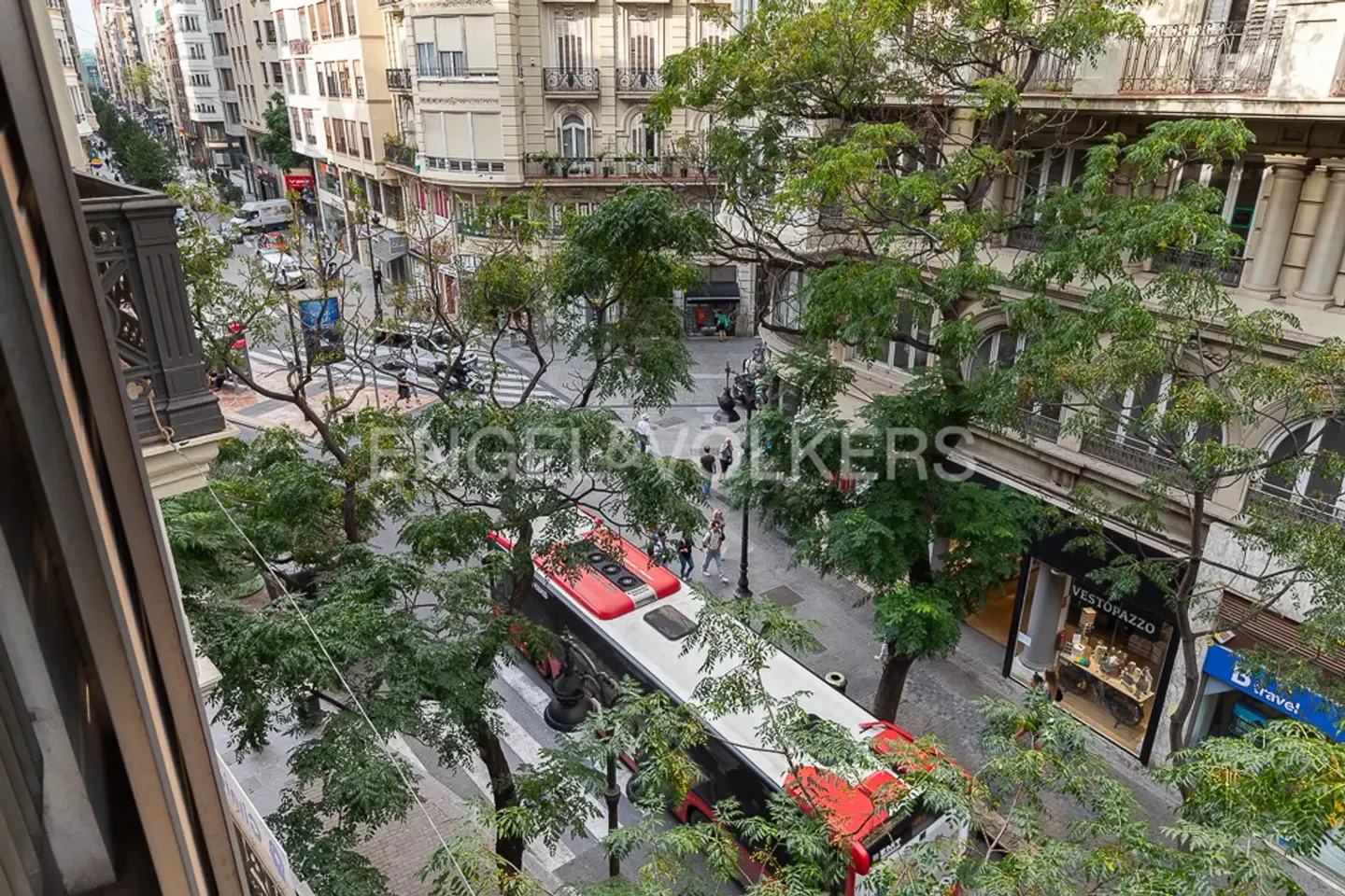 Overhead view of a city street with a red bus, trees, and buildings. People walk on the sidewalks.