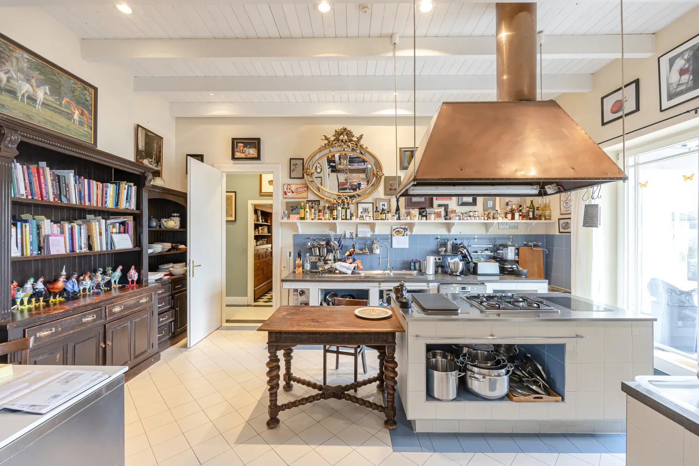 A spacious kitchen with a copper range hood, white cabinets, and a dark wood bookcase filled with books and figurines.