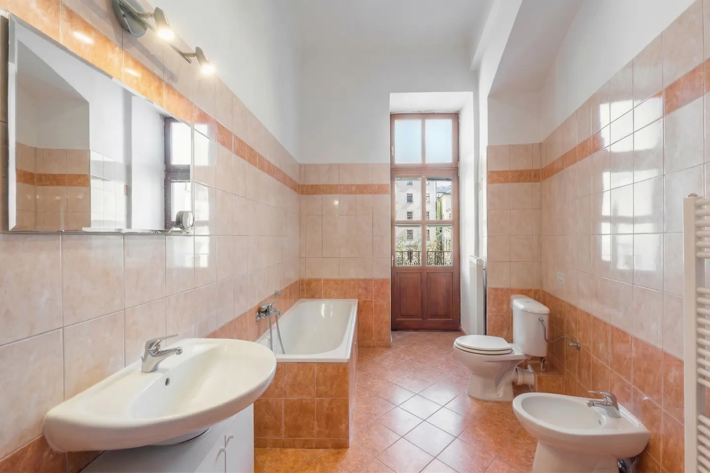 Bathroom with peach-toned tiled walls and floor, featuring a white bathtub, sink, toilet, bidet, and a wooden balcony door.