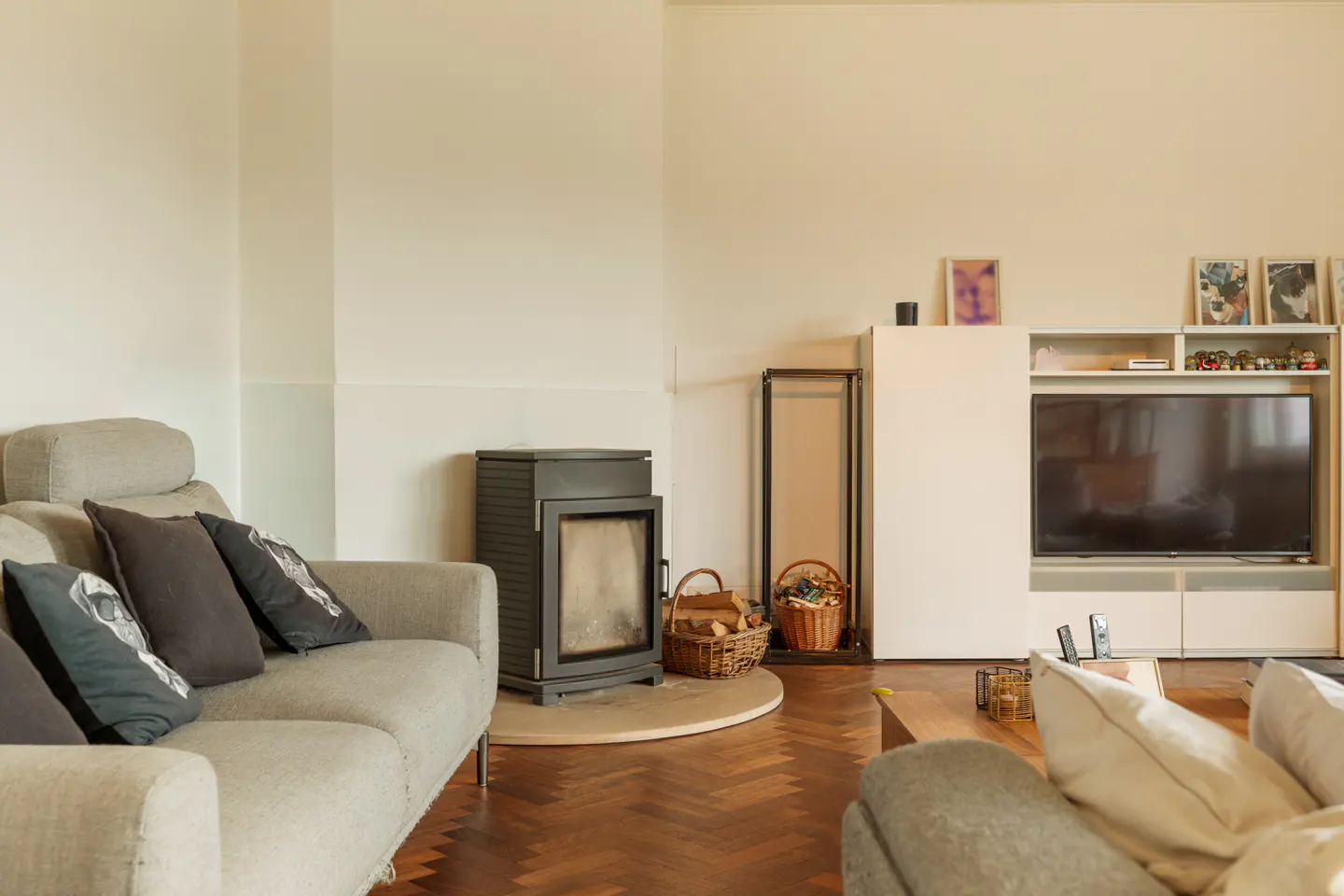 Living room with a grey sofa, wood-burning stove on a stone hearth, television unit, and dark herringbone wood flooring.