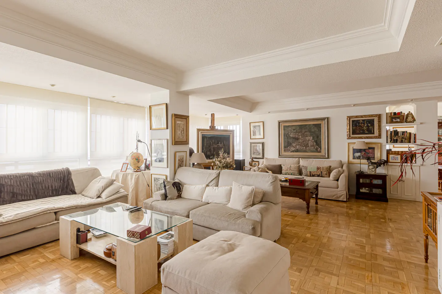Spacious living room with beige sofas, a glass coffee table, parquet floors, and walls decorated with numerous framed art.