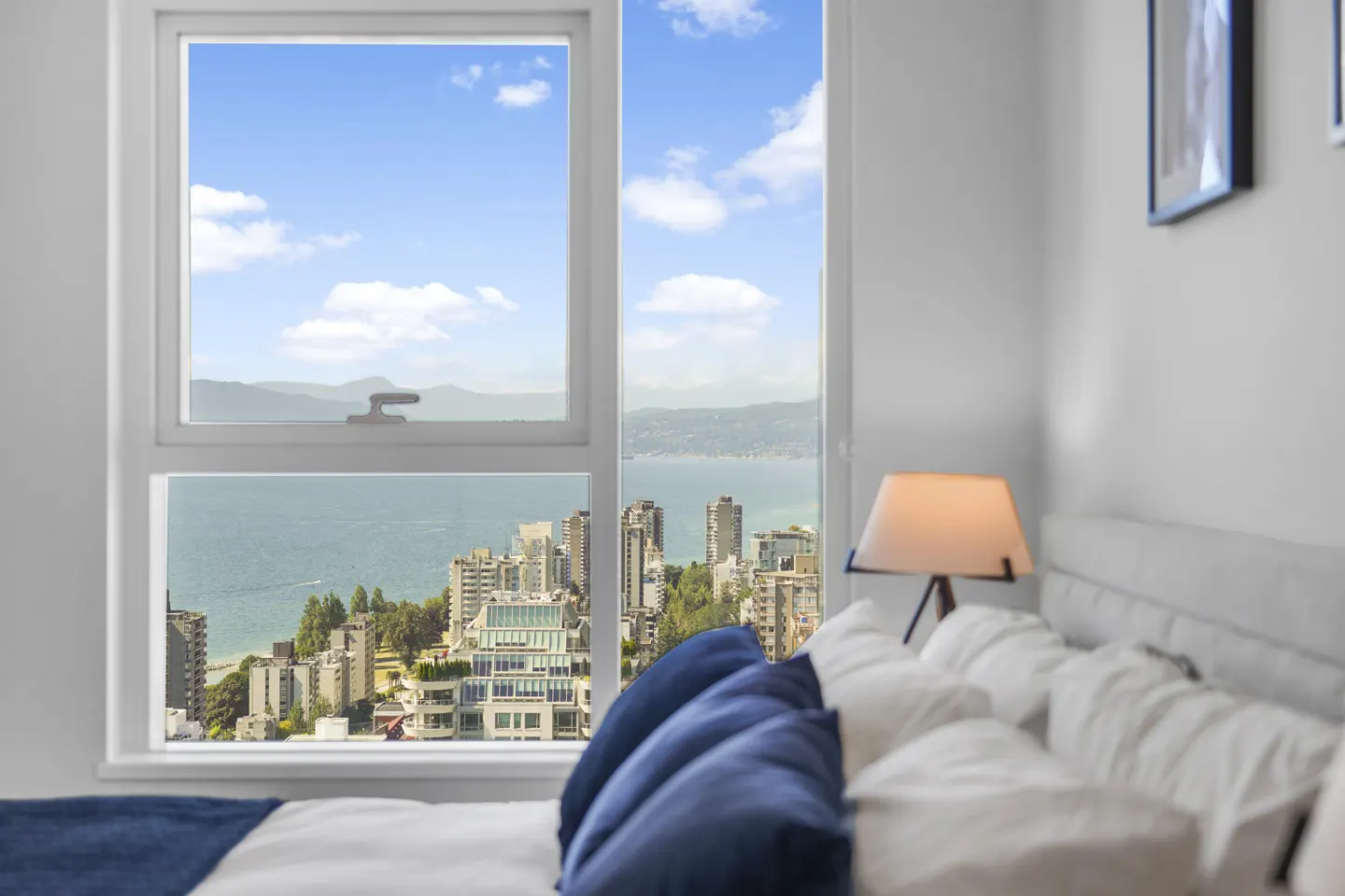 Bedroom view: White bed with blue pillows, lamp, and a large window overlooking a city, ocean, and mountains under a blue sky.
