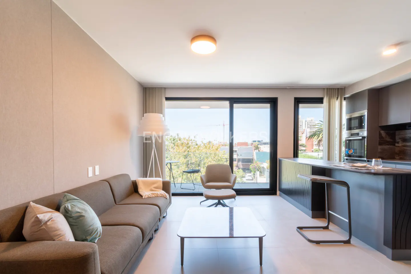 A modern apartment interior with a brown sofa, a coffee table, and a kitchen island. A balcony with a view is visible through the sliding glass doors.