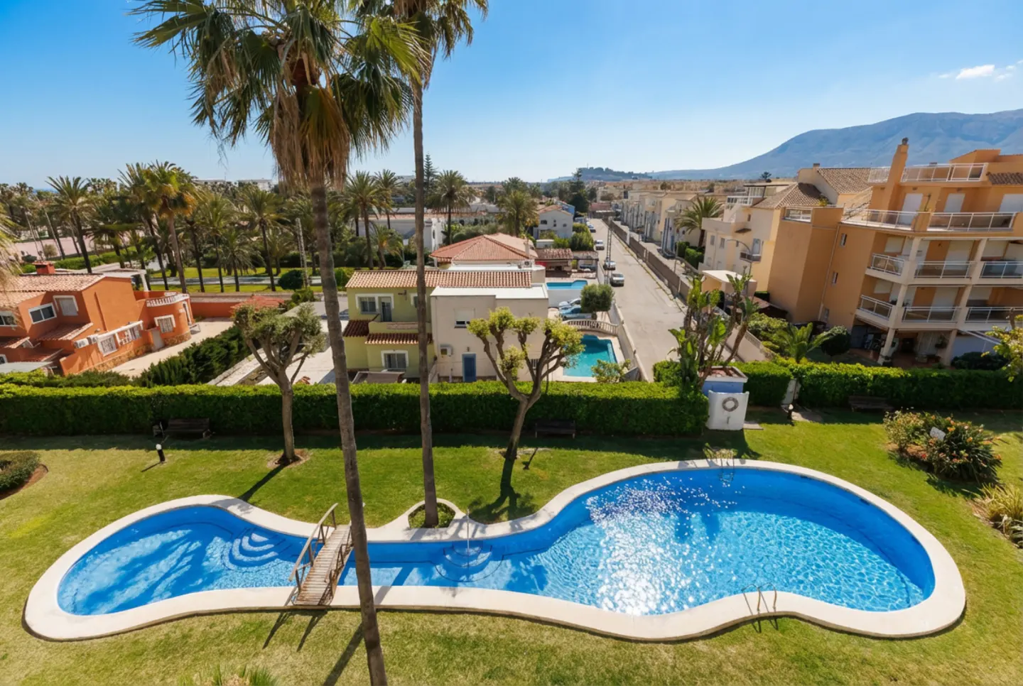 Aerial view of a kidney-shaped pool with a small bridge, surrounded by green grass, palm trees, and residential buildings under a clear blue sky.