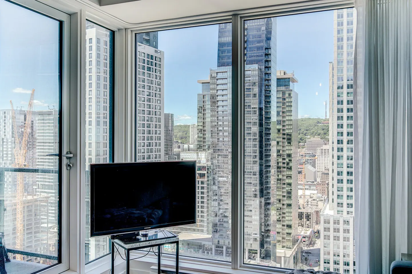 Condo interior with a black TV on a marble table, and a large window view of city skyscrapers on a sunny day.
