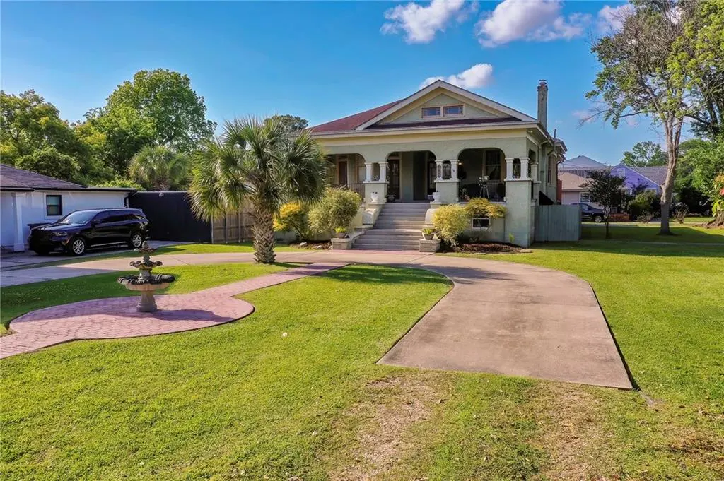 Two-story light green house with a porch, palm tree, fountain, and driveway on a sunny day.