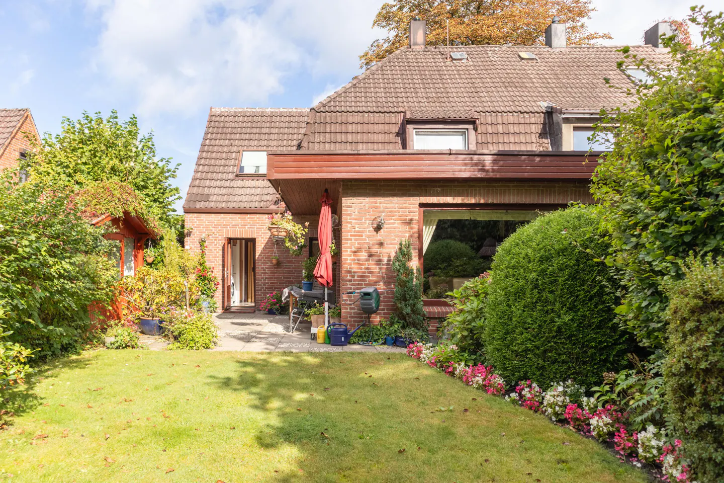 Backyard view of a brick house with a red umbrella, green lawn, and flower beds.