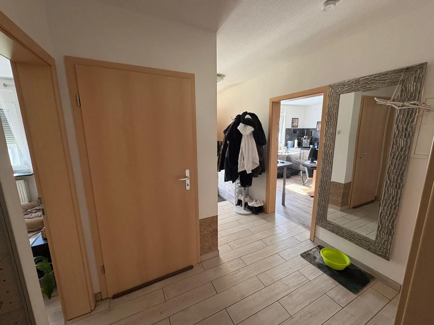 Hallway with light wood doors, tile floor, and a mirror. Coats hang near an open doorway to a kitchen.