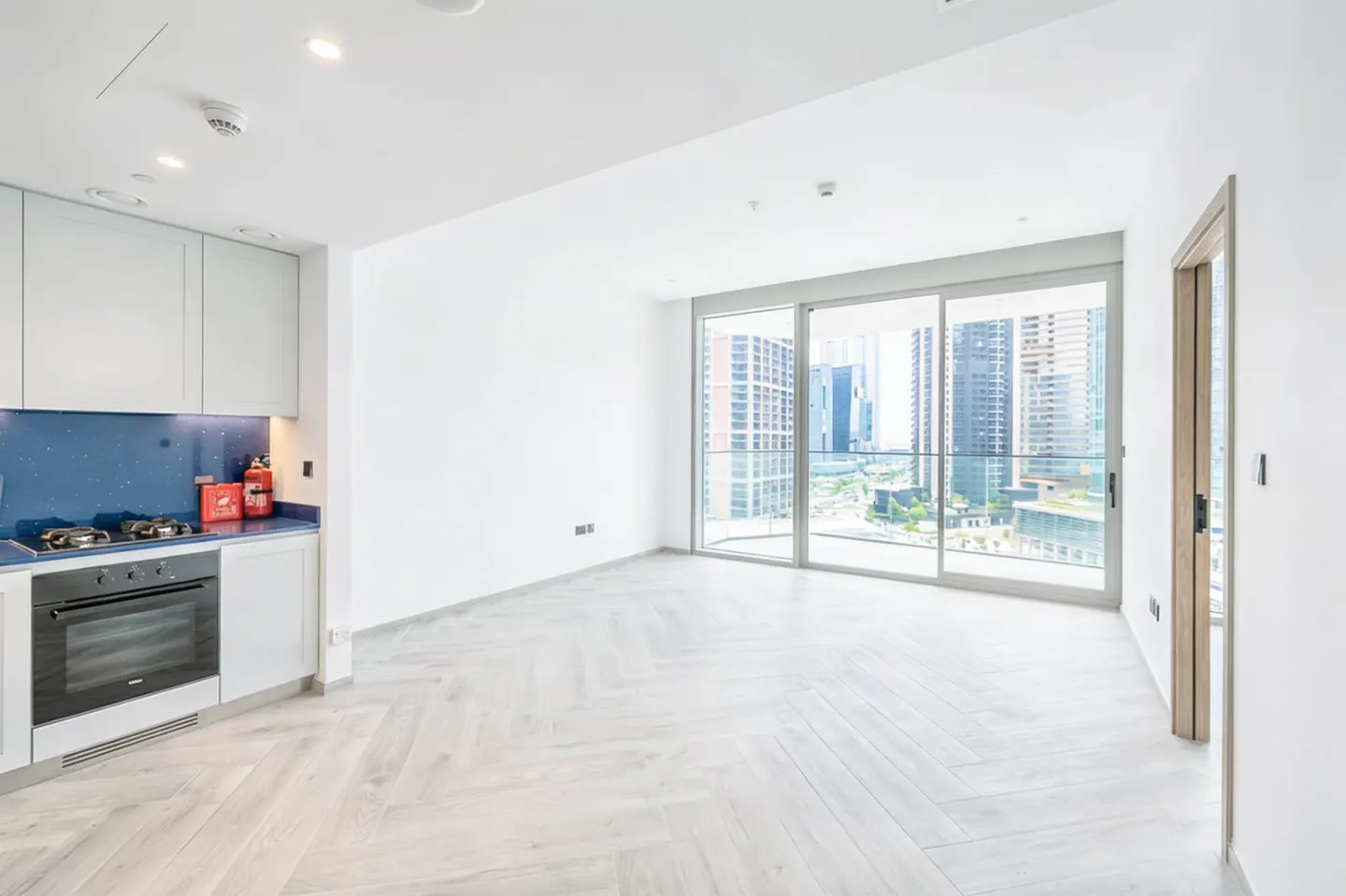 Bright, modern apartment with white walls, herringbone floors, and a kitchen with blue backsplash. Large windows offer a city view.