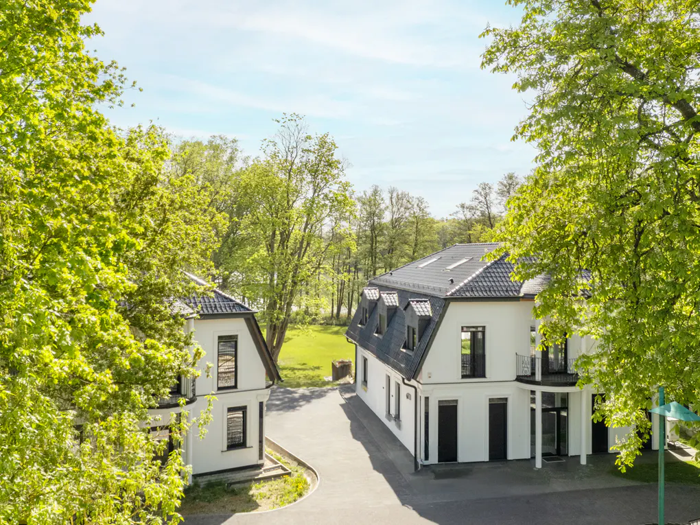 Two modern white houses with black trim and dark roofs sit on a paved driveway, surrounded by green trees and a blue sky.