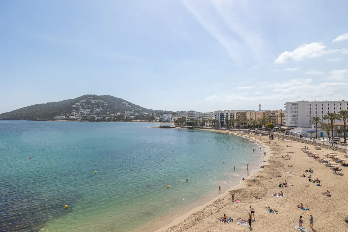 Beach scene with turquoise water, sandy beach, and buildings. People are swimming, sunbathing, and walking along the shore.